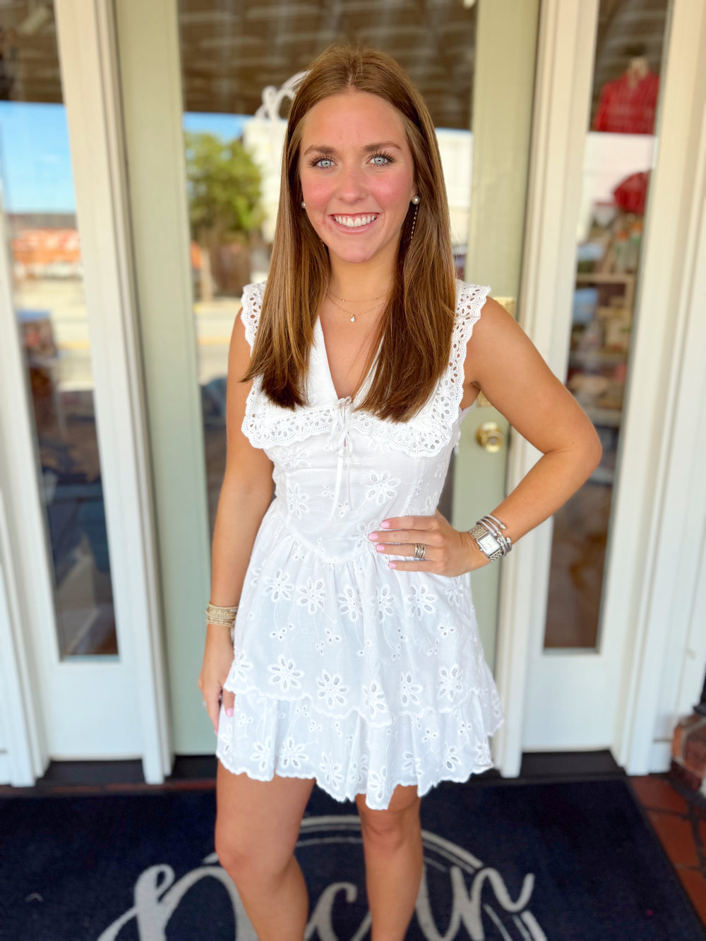 Woman wearing a white floral eyelet mini dress with flutter sleeves, tie-front neckline, scalloped trim, and tiered skirt detail.
