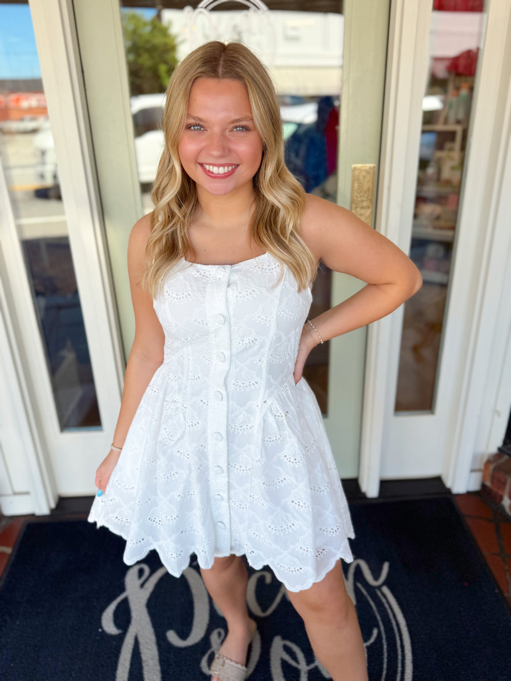 Woman wearing a white embroidered eyelet sleeveless mini dress with button-front detail and scalloped hem.
