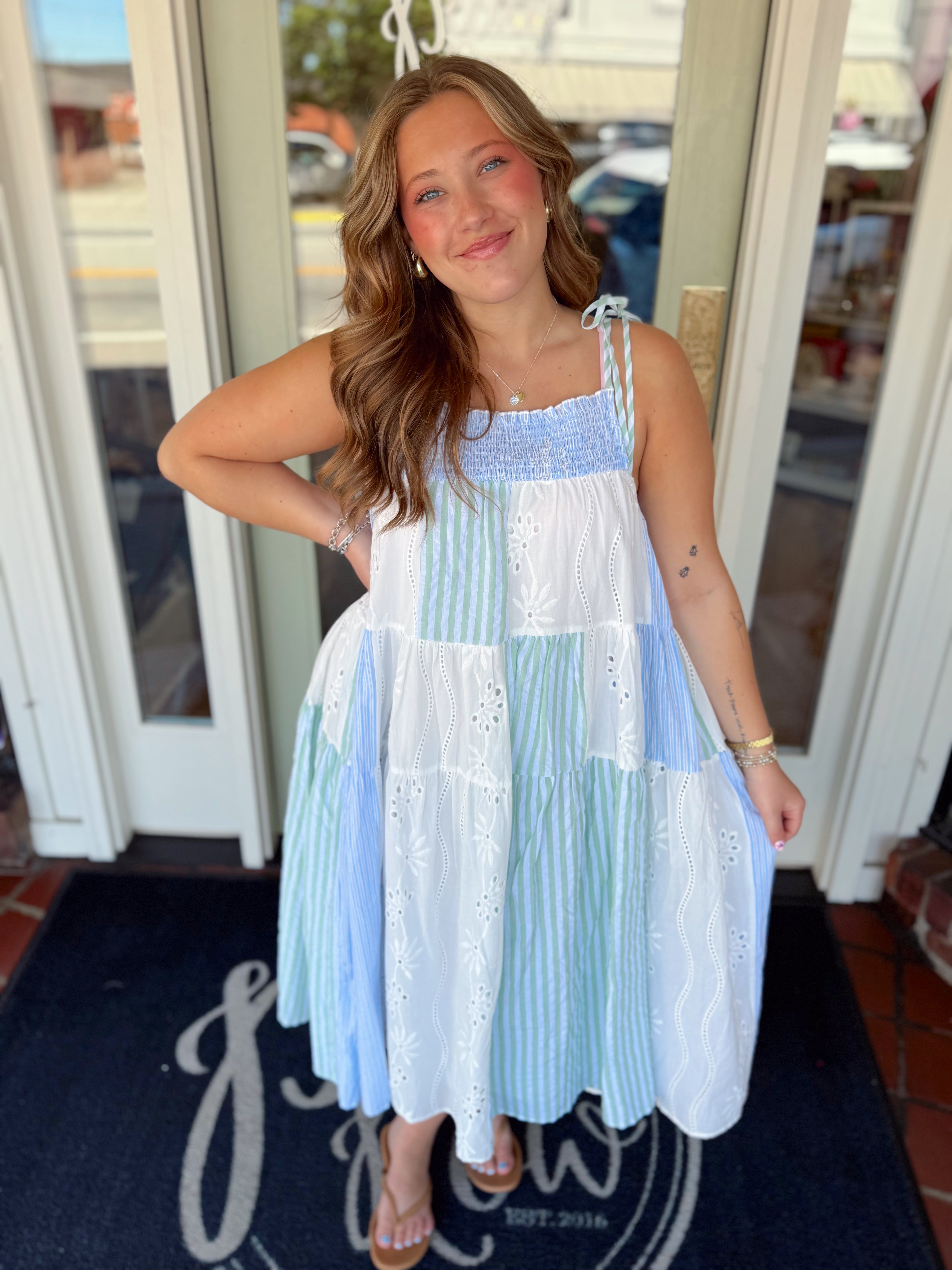 Woman wearing a blue, green, and white patchwork midi dress with striped panels, eyelet embroidery, smocked bodice, and tie shoulder straps.
