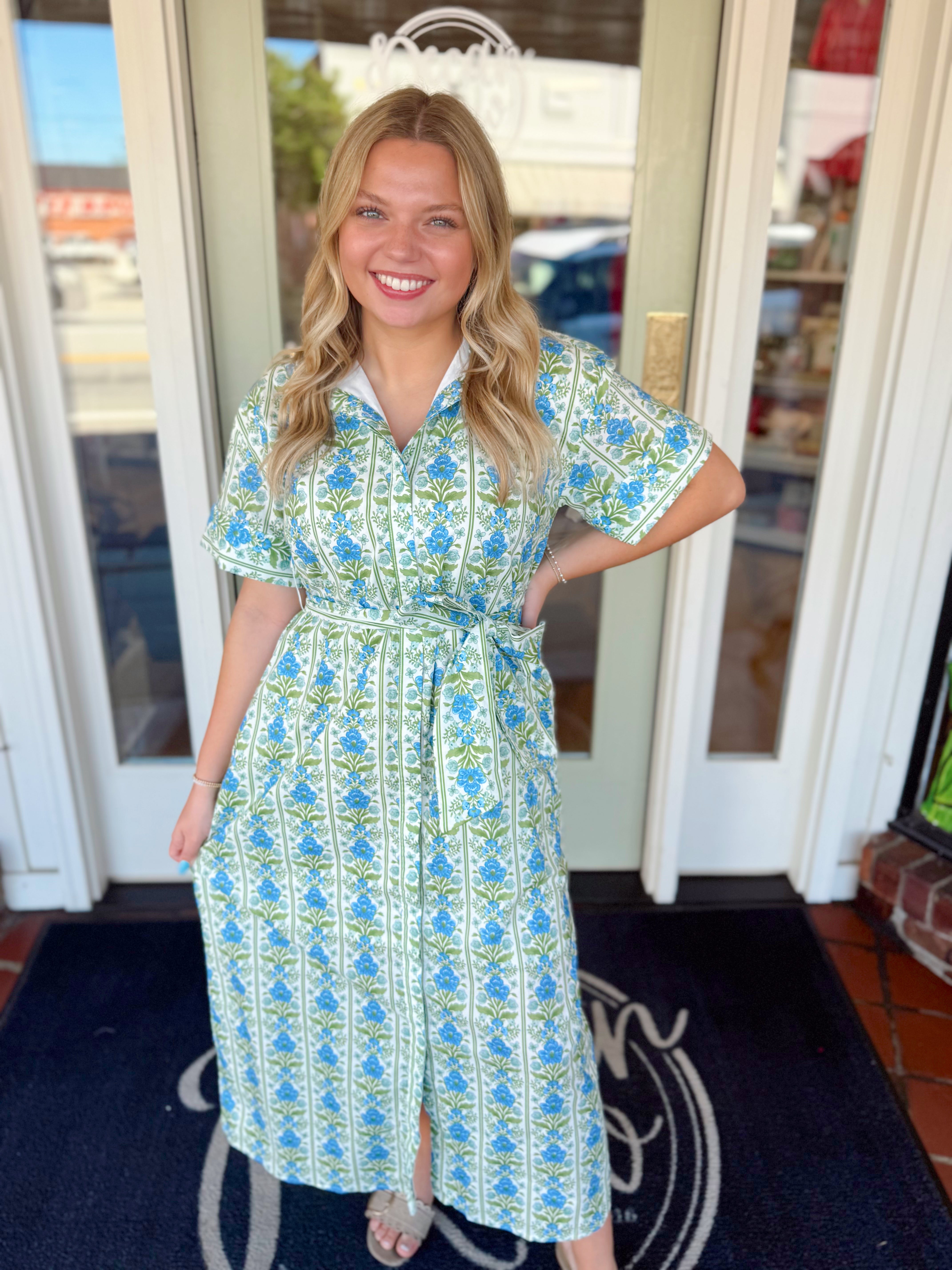Woman wearing a blue and green floral printed midi dress with a collared neckline, short sleeves, and tie waist detail paired with sandals.