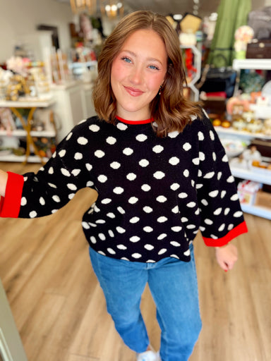  This black knit sweater features an allover cream polka dot pattern with bold red contrast trim at the neckline and cuffs. 