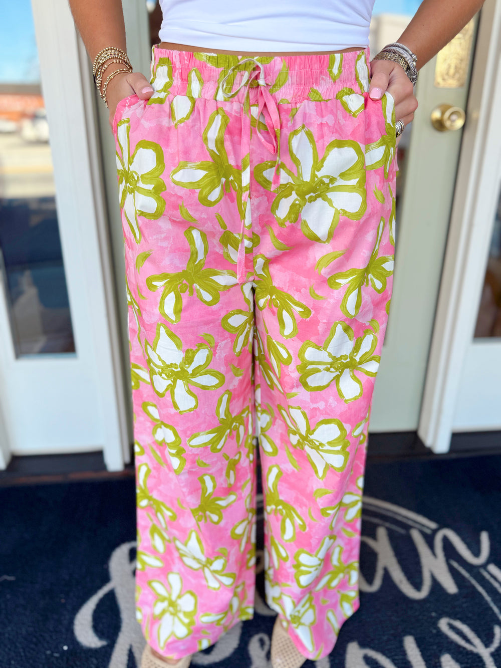 Pink wide-leg floral pants with lime green and white oversized flower print, elastic drawstring waist, and flowy relaxed fit.