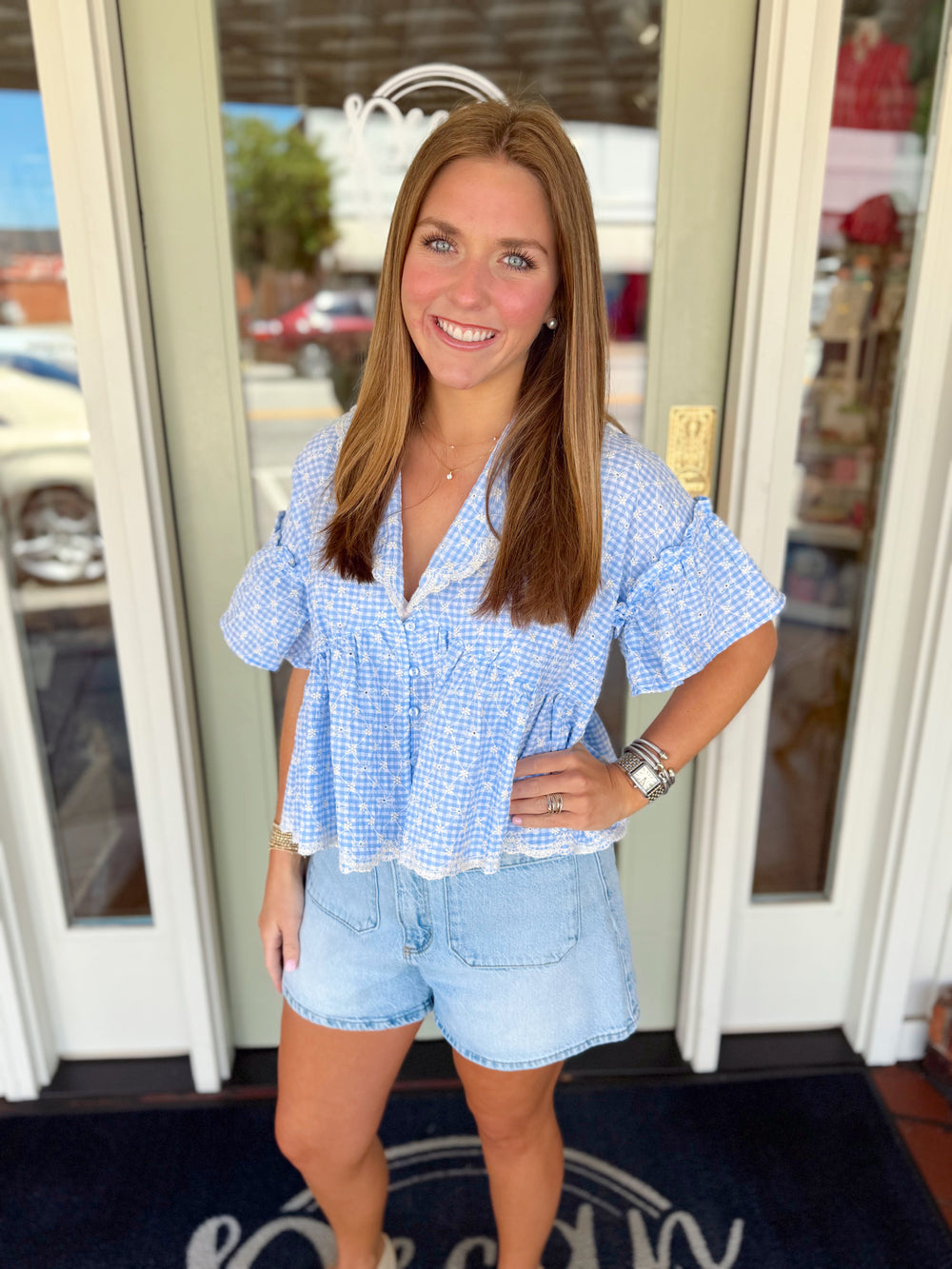 Woman wearing a blue and white gingham blouse with flutter sleeves and eyelet embroidery detail paired with light wash denim shorts.

