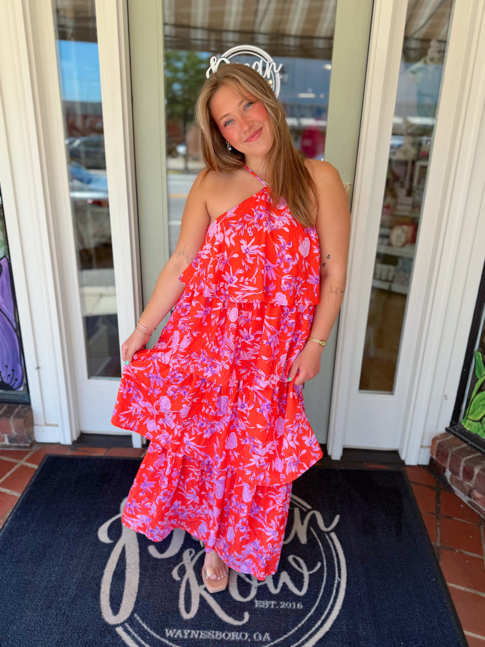 Coral tiered maxi dress featuring a halter neckline and an allover floral print in shades of lavender and soft pink, styled with neutral sandals for a bright summer look.