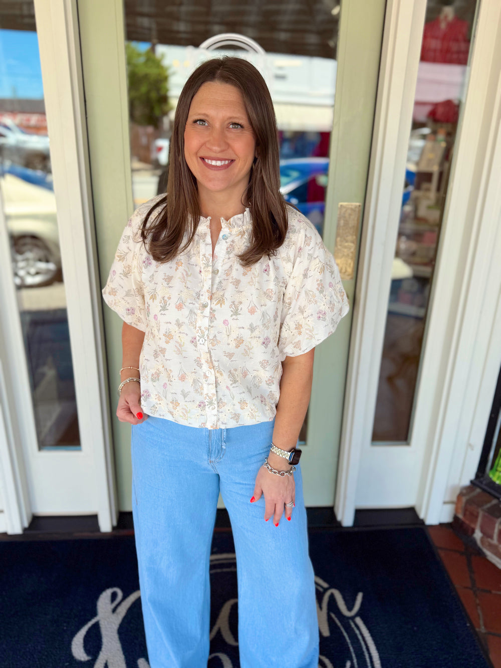 Woman wearing a cream floral print button-down blouse with short sleeves and ruffle neckline detail paired with light wash denim jeans.