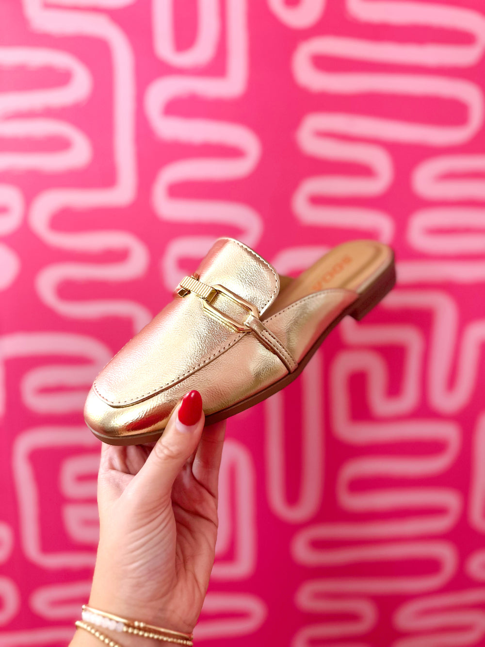 A metallic gold slip-on loafer with a gold buckle detail, held by a hand with red nail polish against a vibrant pink background with white abstract lines.