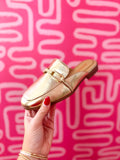 A metallic gold slip-on loafer with a gold buckle detail, held by a hand with red nail polish against a vibrant pink background with white abstract lines.