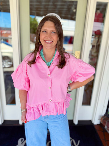 Woman wearing a bright pink button-down blouse with puff sleeves and ruffle hem detail paired with light wash denim jeans and a turquoise layered necklace.
