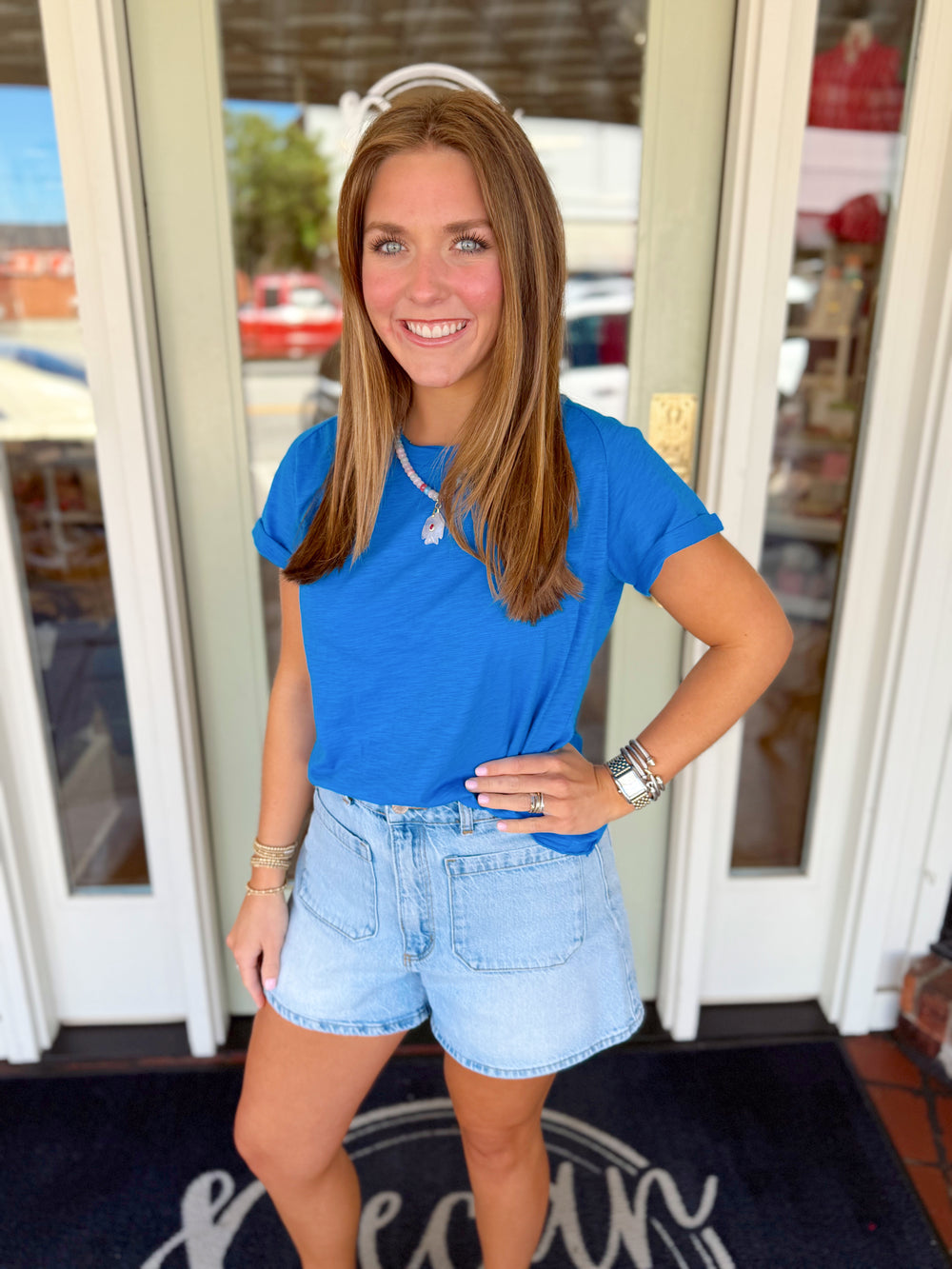 Woman wearing a royal blue short sleeve tee paired with light wash denim shorts and layered jewelry. 