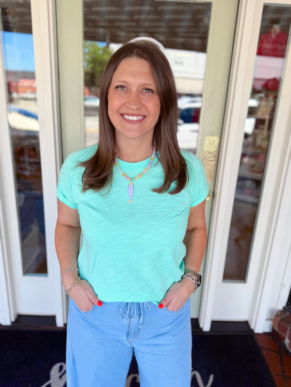 Woman wearing a mint green short sleeve tee paired with light wash denim jeans and a colorful beaded necklace.
