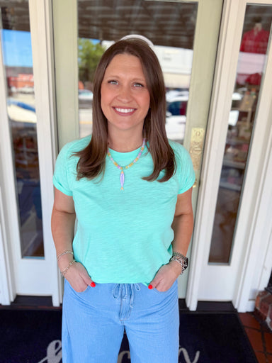 Woman wearing a mint green short sleeve tee paired with light wash denim jeans and a colorful beaded necklace.
