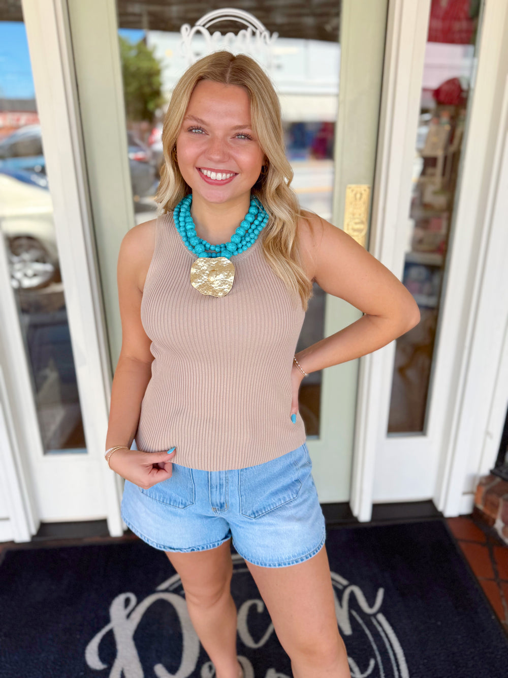 Woman wearing a taupe ribbed sleeveless knit tank top paired with light wash denim shorts and a turquoise statement necklace.
