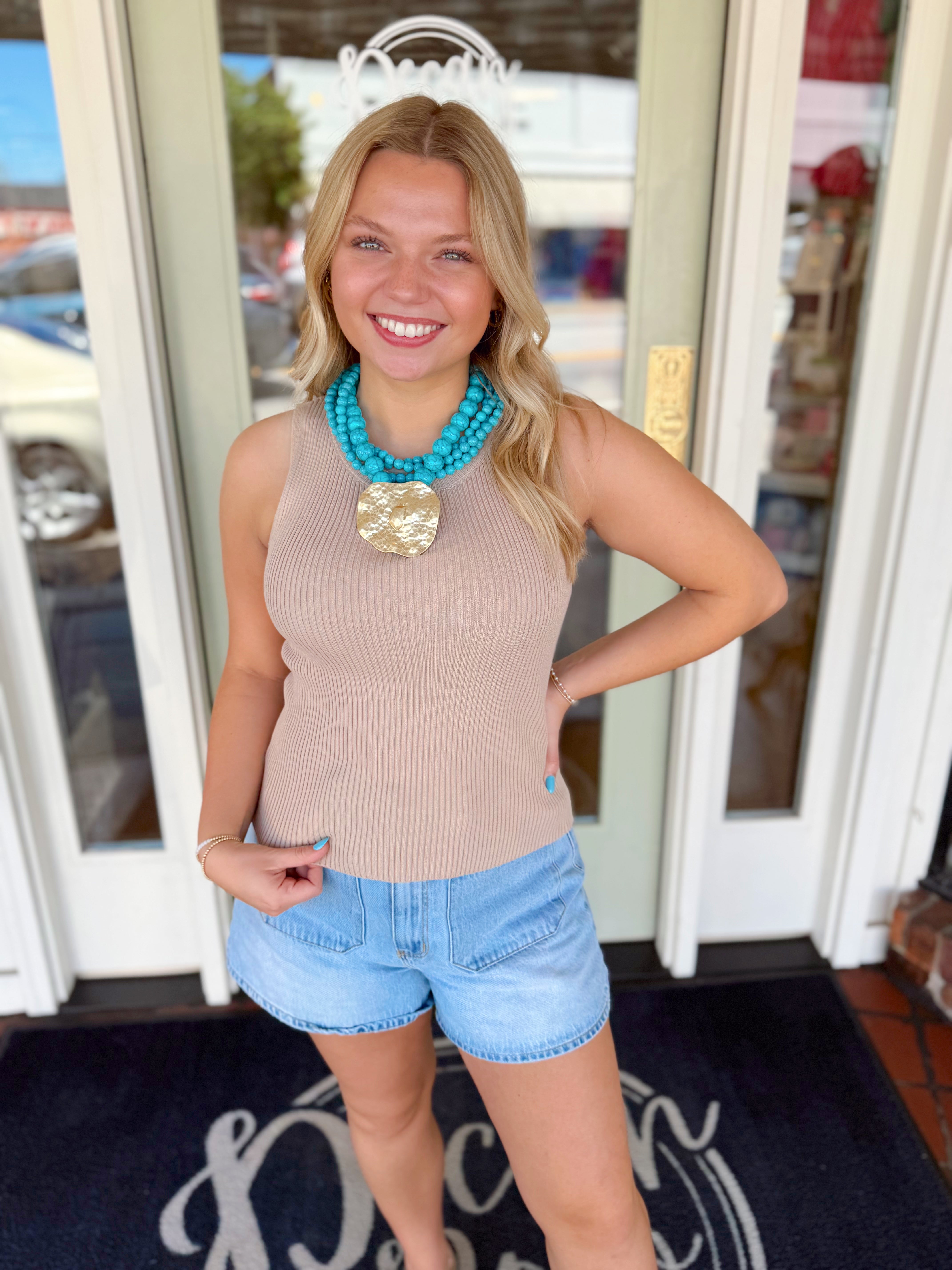 Woman wearing a taupe ribbed sleeveless knit tank top paired with light wash denim shorts and a turquoise statement necklace.
