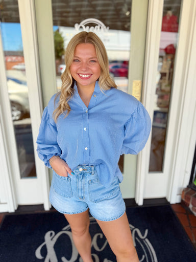 Woman wearing a blue striped button-down blouse with oversized puff sleeves paired with light wash denim shorts.
