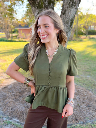 This olive top features a flattering V-neckline with fabric-covered buttons, softly puffed short sleeves, and a gathered peplum waist that gives it just the right amount of shape.