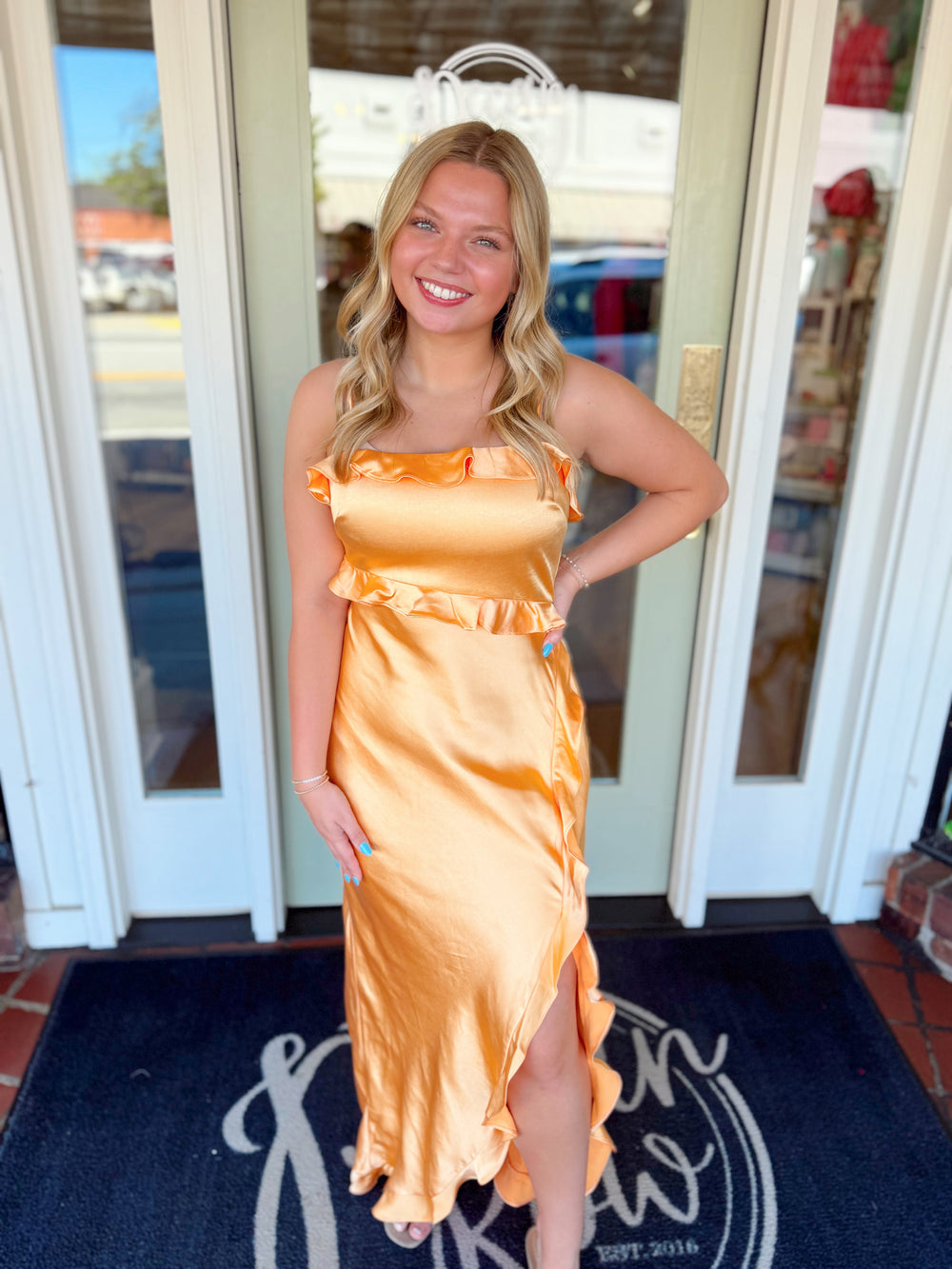 Woman wearing an orange satin midi dress with ruffle trim details and asymmetrical hem.
