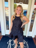 Woman wearing a sleeveless black floral lace mini dress with a ruffle hem and relaxed silhouette.
