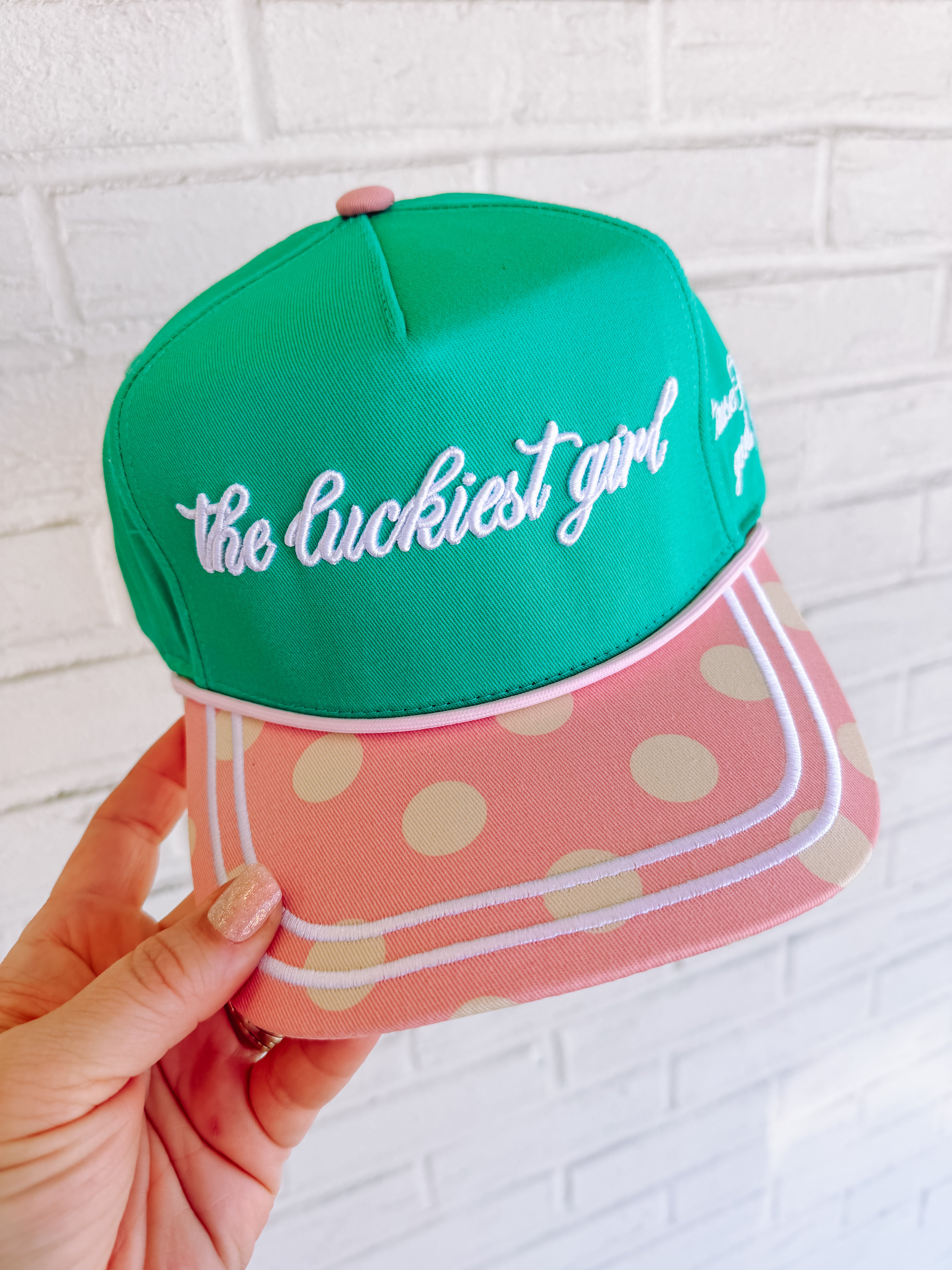 Green cap with 'the luckiest girl' text held by a hand against a white brick wall.