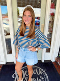Woman wearing a blue and taupe striped oversized top with wide sleeves paired with light wash denim shorts.
