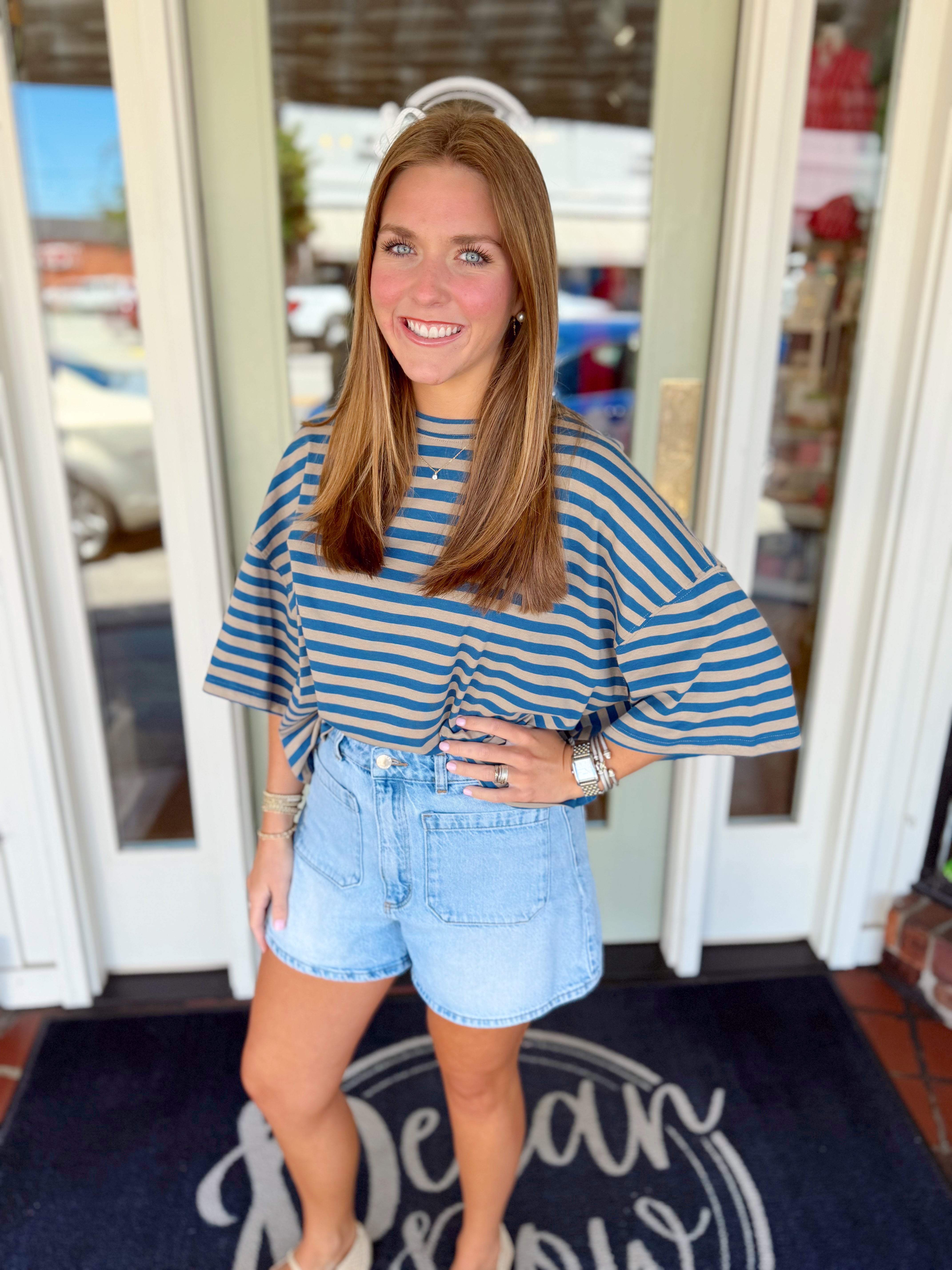 Woman wearing a blue and taupe striped oversized top with wide sleeves paired with light wash denim shorts.
