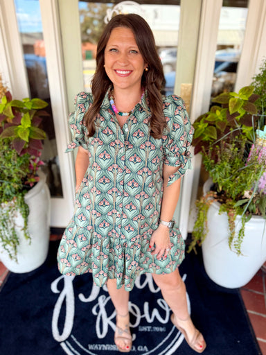 This green button-front dress features an allover botanical floral print in soft, earthy tones, a classic collared neckline and subtle puff sleeves with tie details