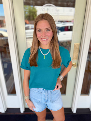 Woman wearing a deep teal short sleeve tee paired with light wash denim shorts and a white beaded necklace with green pendant detail.
