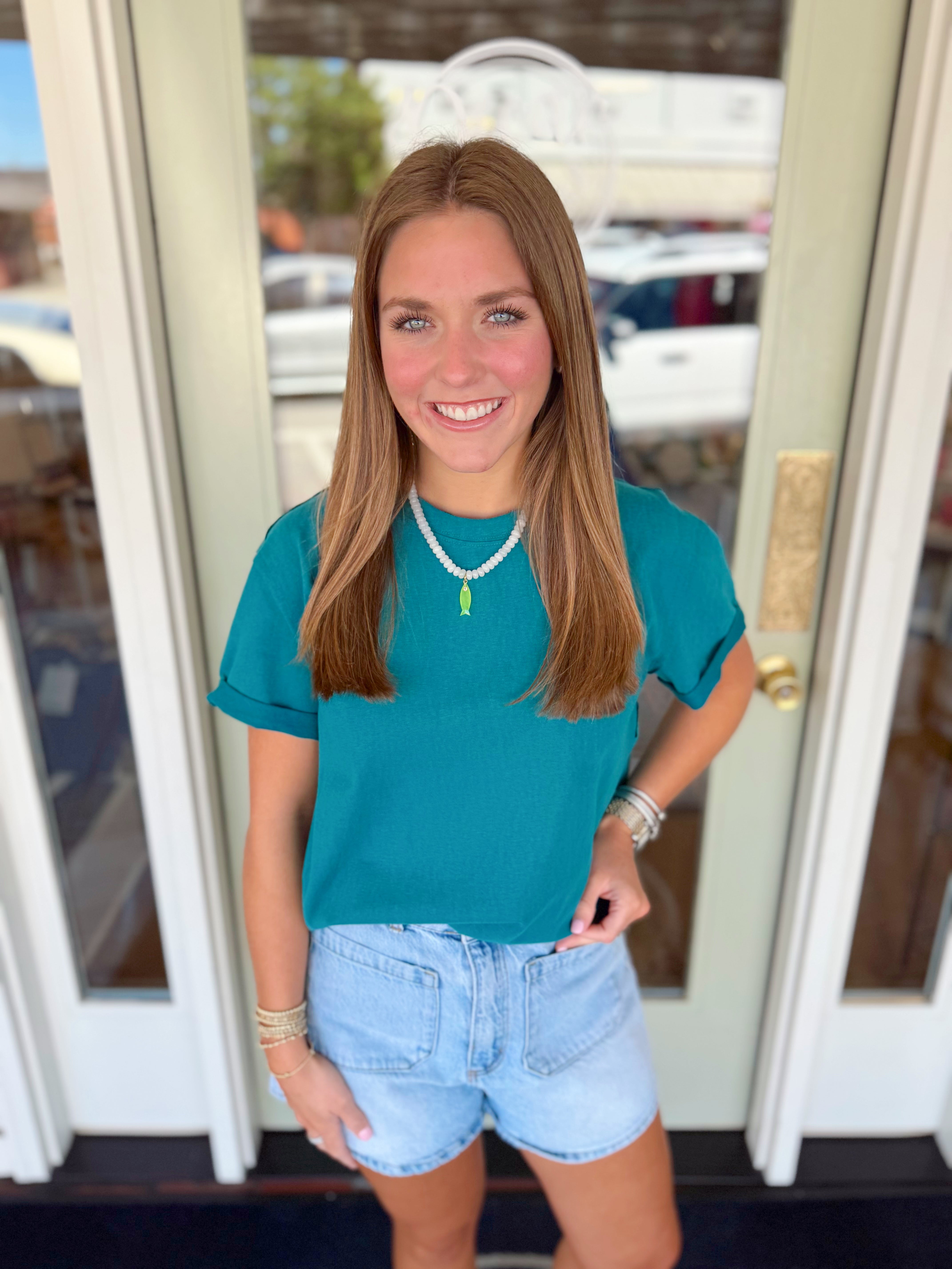Woman wearing a deep teal short sleeve tee paired with light wash denim shorts and a white beaded necklace with green pendant detail.
