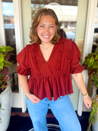This rich rust blouse features a flattering V-neckline framed with delicate ruffle trim, puff sleeves and a gathered peplum hem.