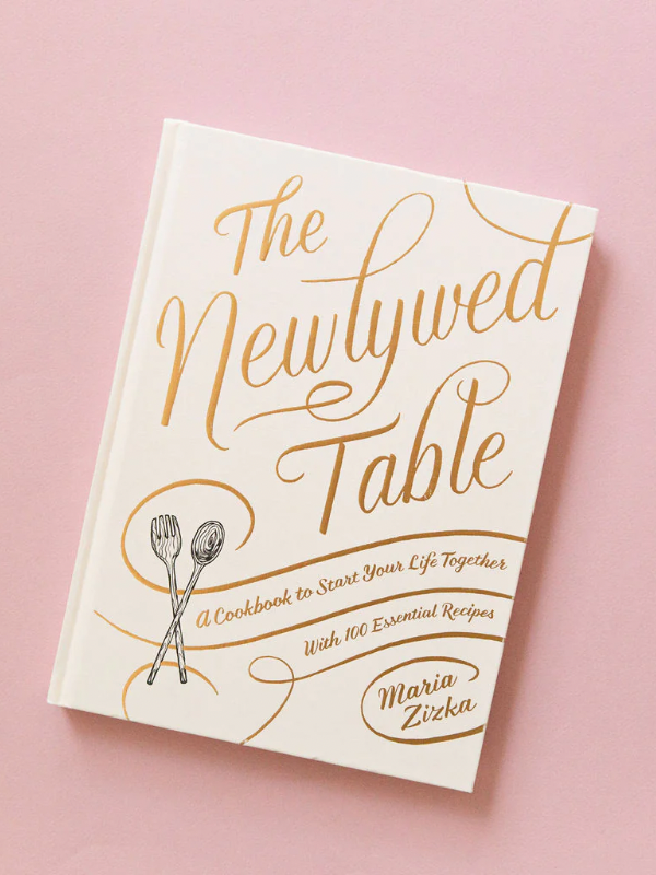 A cookbook titled 'The Newlywed Table' by Maria Zizka, featuring a white cover with gold lettering and illustrations of crossed spoons.