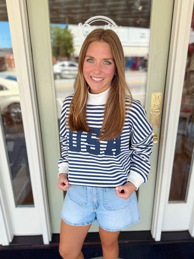 Woman wearing a navy and white striped USA pullover sweatshirt paired with light wash denim shorts.
