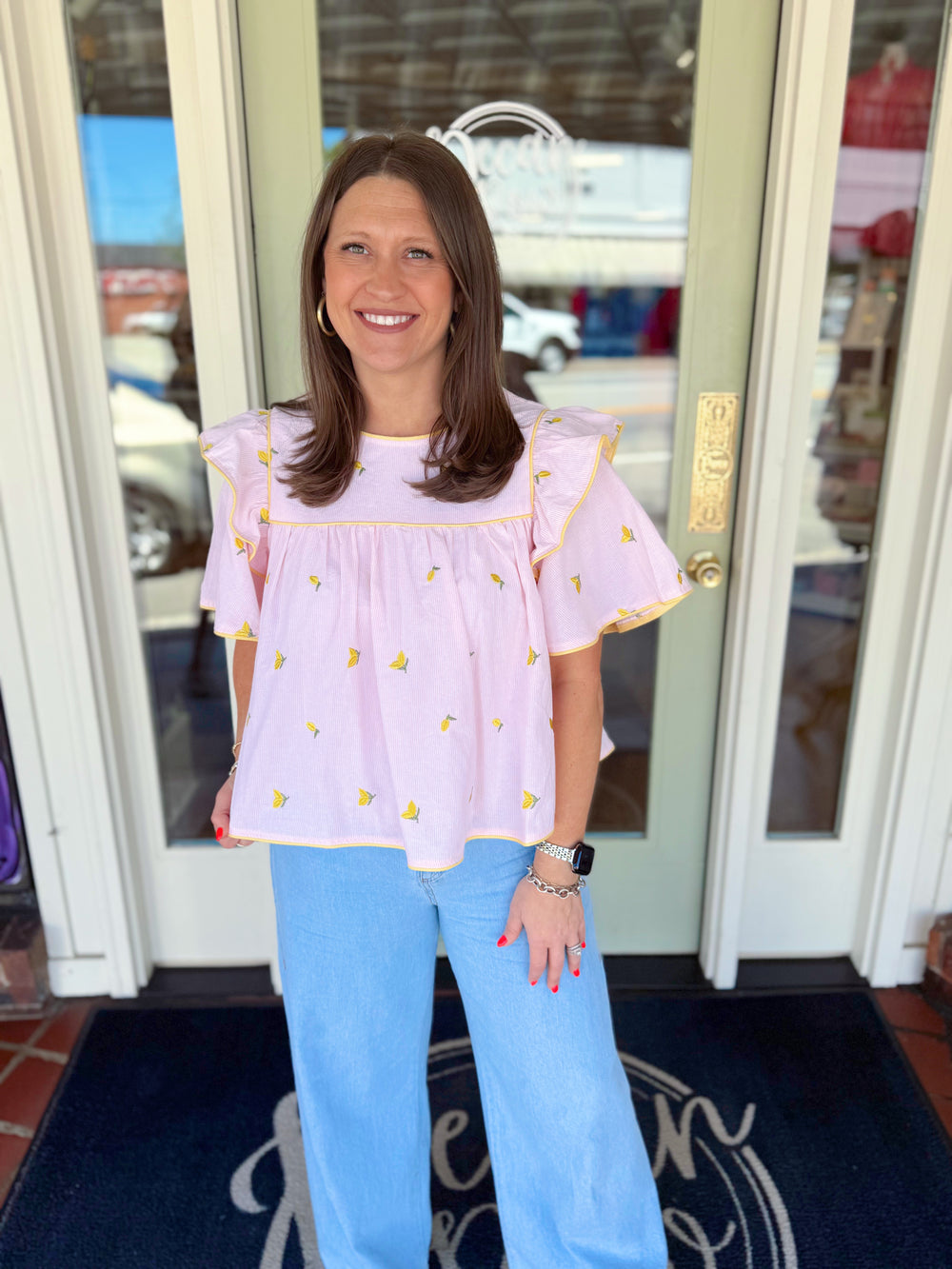 Woman wearing a lavender embroidered blouse with yellow floral details and flutter sleeves paired with light wash denim jeans.
