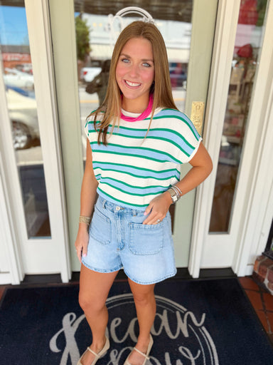 Woman wearing an ivory knit top with green and blue horizontal stripes paired with light wash denim shorts and a pink statement necklace.
