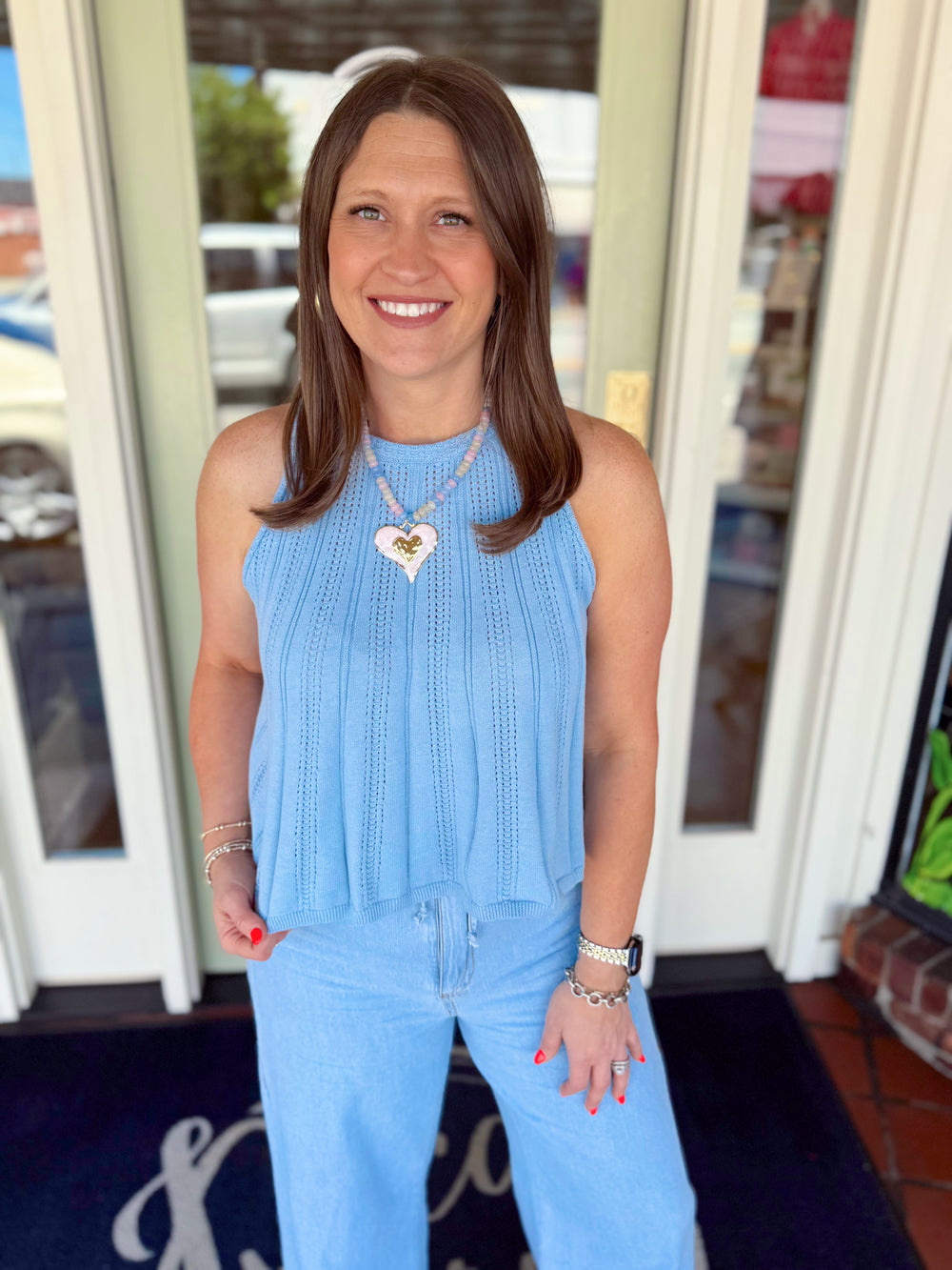 Woman wearing a soft blue sleeveless pointelle knit sweater tank paired with light wash denim jeans and a heart pendant necklace.