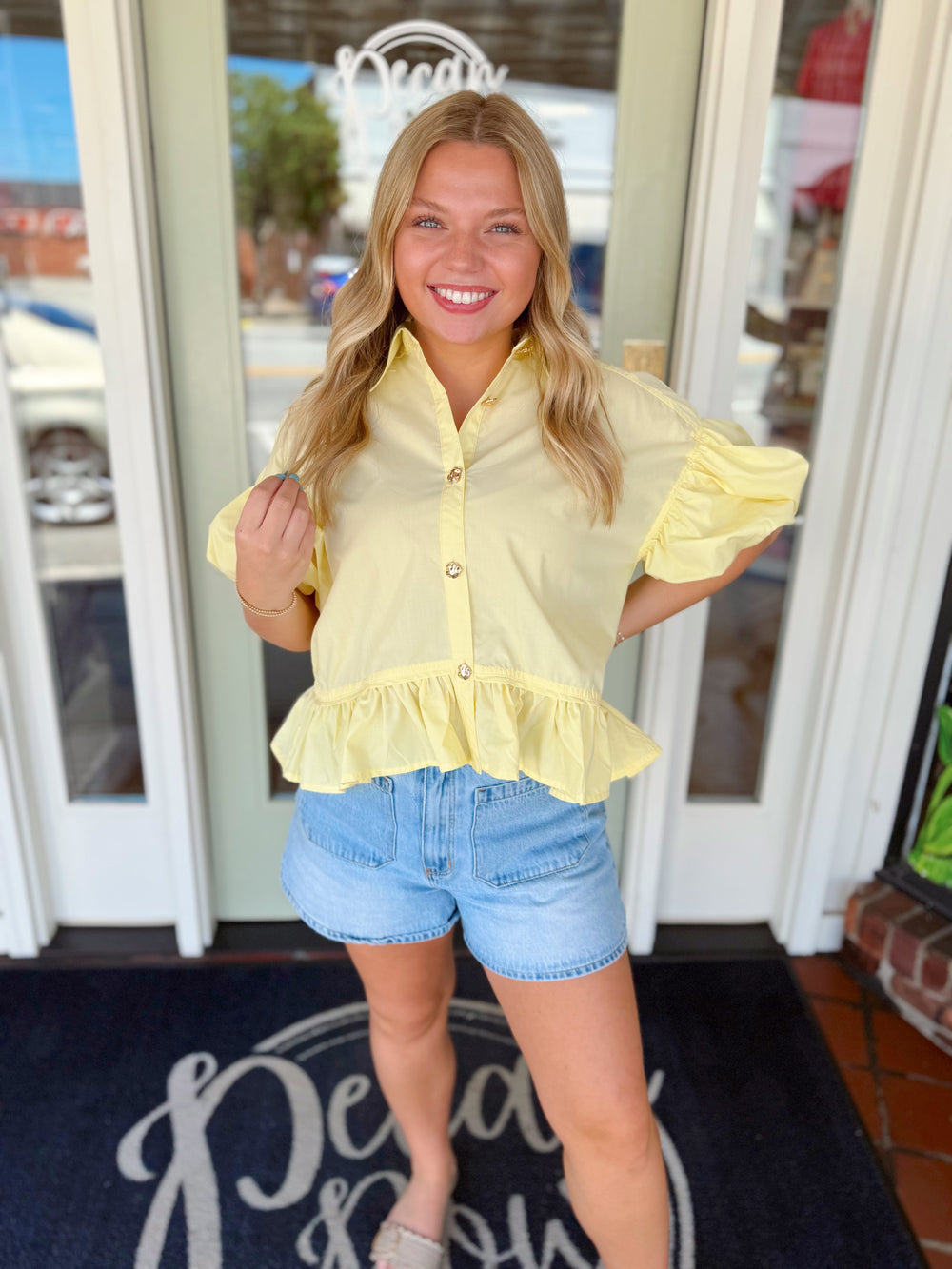 Woman wearing a buttery yellow puff sleeve peplum top with gold buttons and ruffle hem paired with light wash denim shorts.
