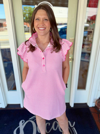 Woman wearing a pink and white gingham mini dress with scalloped flutter sleeves, collared neckline, button detail, and side pockets.