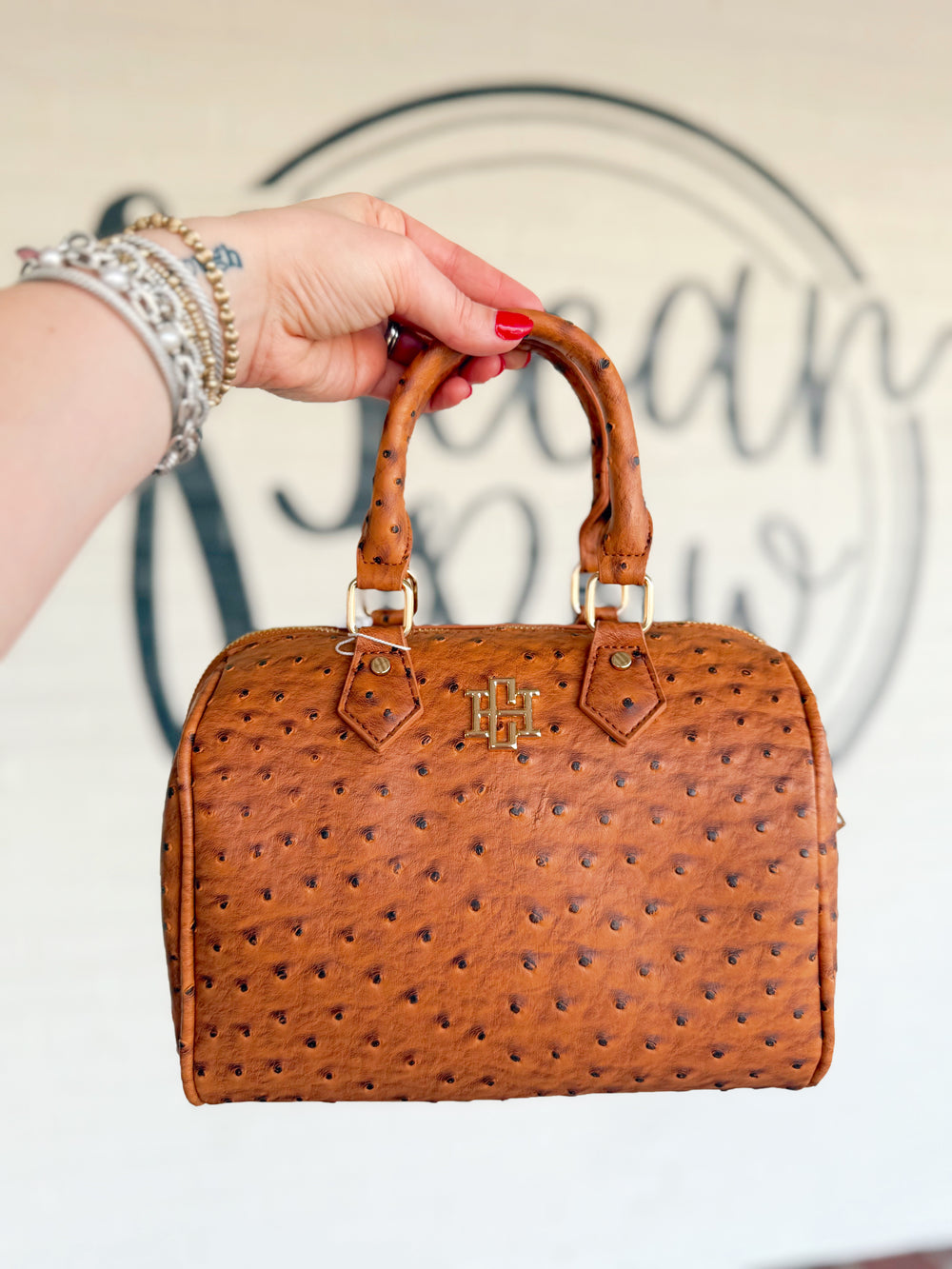 This rich cognac handbag features an ostrich-inspired textured finish, structured barrel silhouette, and coordinating top handles.
