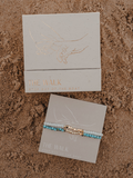 Turquoise and gold bracelet on a branded card with 'The Walk' packaging on sand.