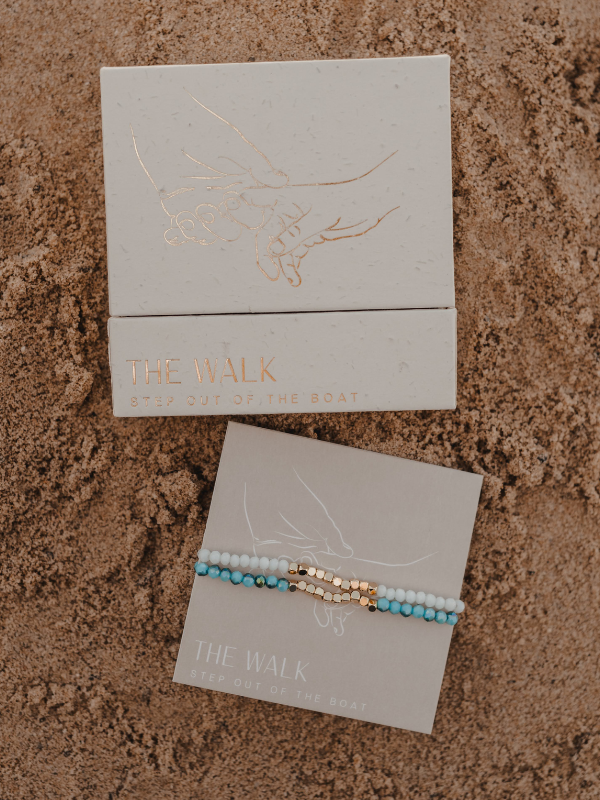 Turquoise and gold bracelet on a branded card with 'The Walk' packaging on sand.