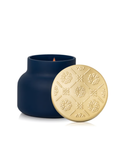 A dark navy blue candle jar with its embossed floral gold lid leaning against it. 