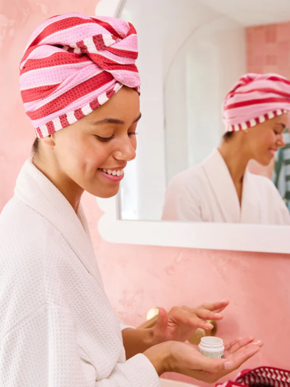 Woman applying cream in front of a mirror wearing a pink and white striped towel on her head.