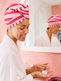 Woman applying cream in front of a mirror wearing a pink and white striped towel on her head.