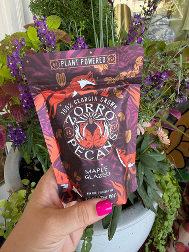 A hand with bright pink nail polish holds a bag of Zorro Pecans Maple Glazed peanuts in front of a potted plant with purple flowers and green foliage.