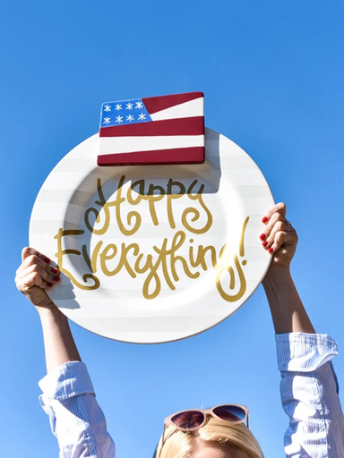 Person holding a plate with 'Happy Everything!' and an American flag against a blue sky.
