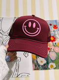 Maroon cap with pink smiley face held against a colorful and patterned background