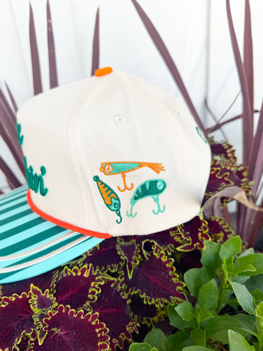 Cream trucker hat with embroidered “Gone Fishin’” lettering, aqua striped brim, fishing lure embroidery on the side, and “Catch Ya Later” embroidered on the back.
