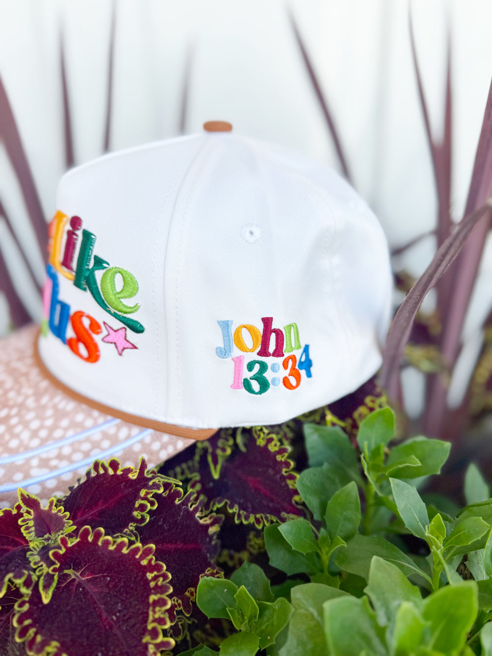 White trucker hat with colorful embroidered “Love Like Jesus” lettering, spotted tan brim, John 13:34 side embroidery, and colorful faith-inspired embroidered icons on the back.
