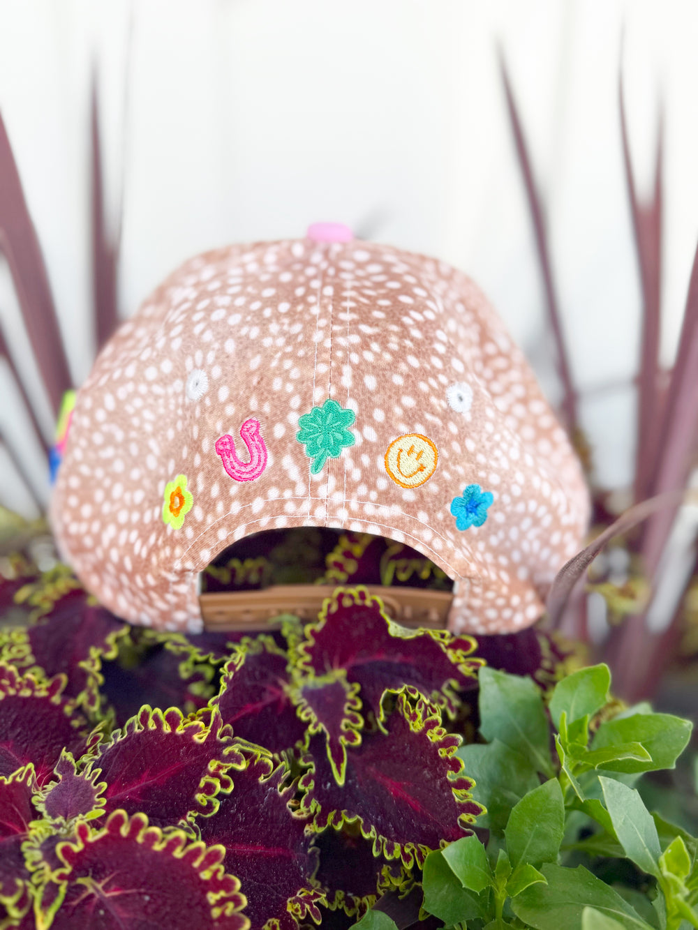 Tan spotted trucker hat with colorful embroidered “Make It a Good Day” lettering, “Just Be Happy” side embroidery, and multicolor lucky-inspired embroidered icons on the back.

