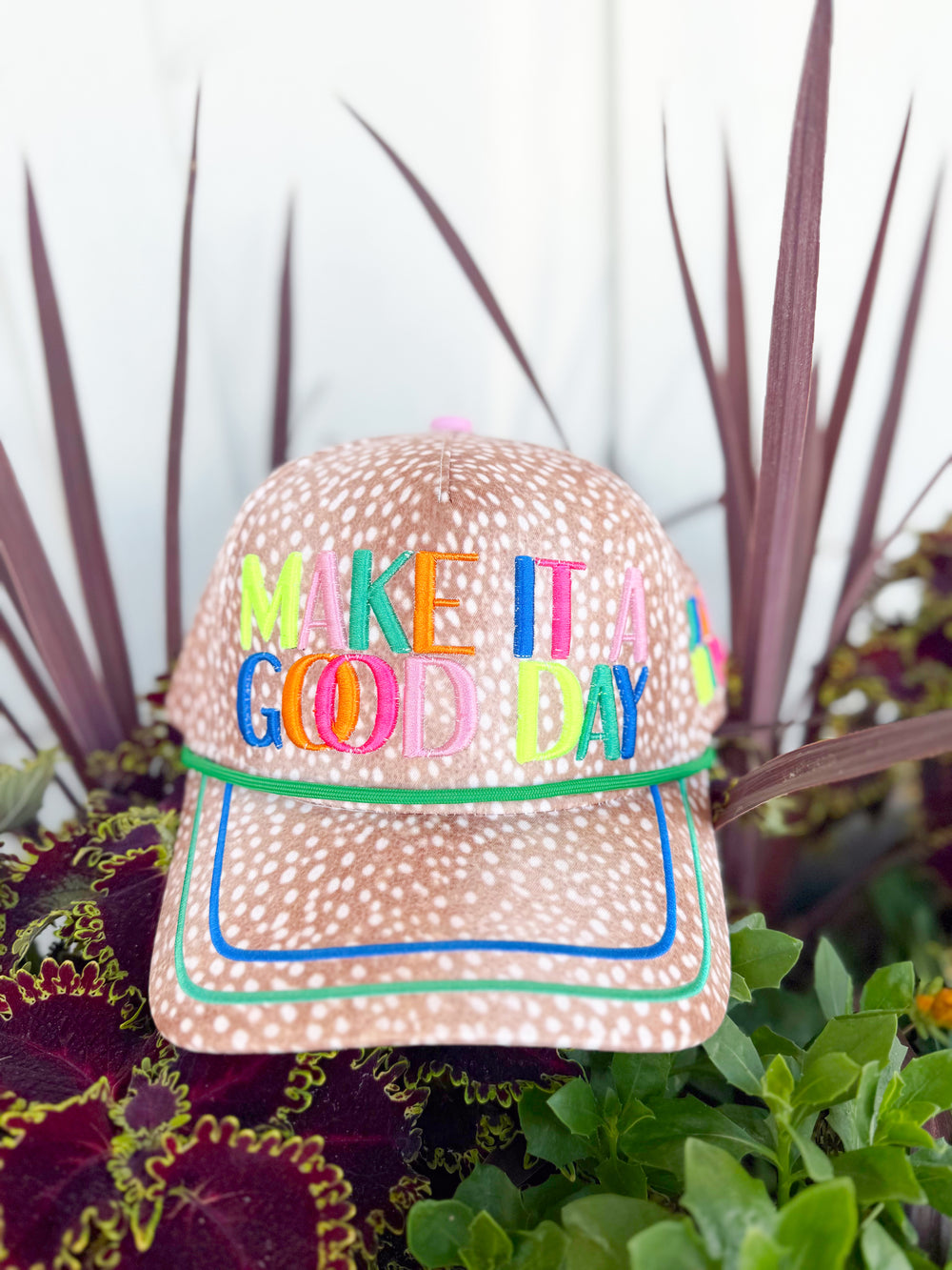 Tan spotted trucker hat with colorful embroidered “Make It a Good Day” lettering, “Just Be Happy” side embroidery, and multicolor lucky-inspired embroidered icons on the back.
