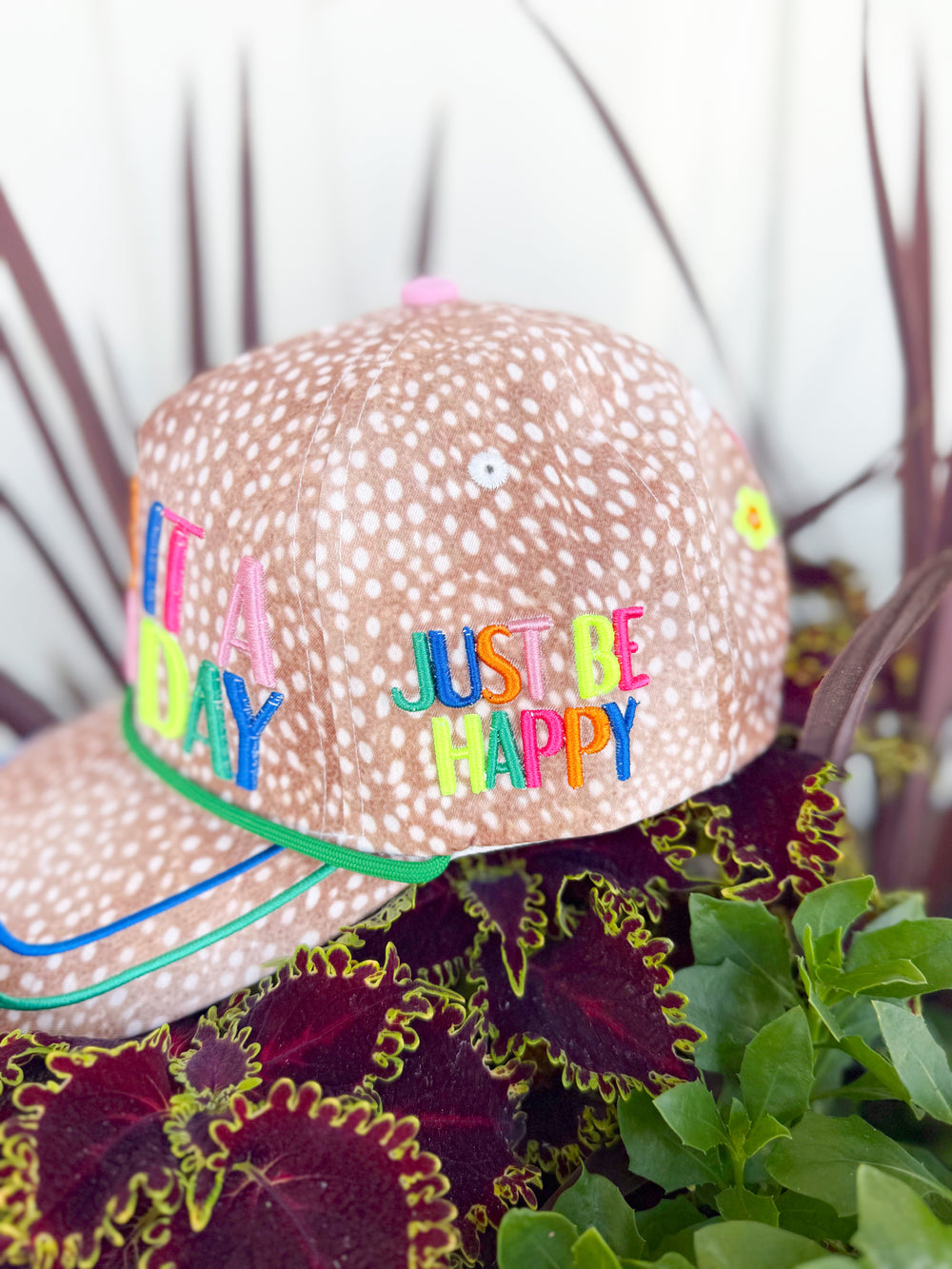 Tan spotted trucker hat with colorful embroidered “Make It a Good Day” lettering, “Just Be Happy” side embroidery, and multicolor lucky-inspired embroidered icons on the back.
