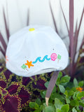 White trucker hat with colorful “It’s a Good Day” embroidery, rainbow striped brim, and beach-themed embroidered details including palm trees, waves, seashells, and starfish.
